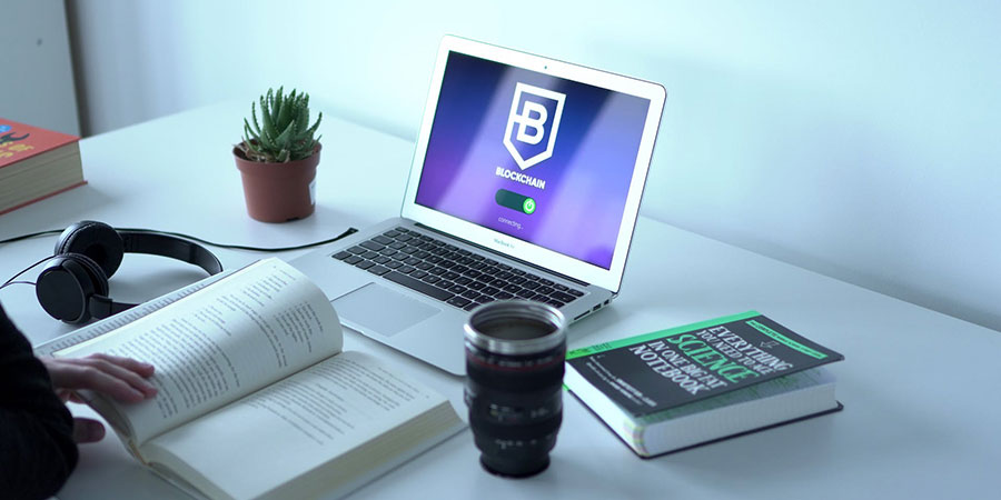 close up view of a person reading a book in front of a macbook air with headphones, a plant, a book and a cup on top of the table
